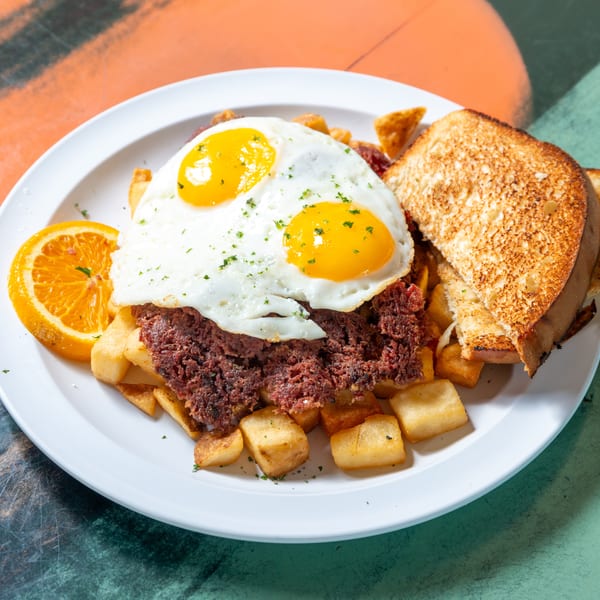 Corned beef hash with two fried eggs, home fries, toast, and an orange slice on a white plate.