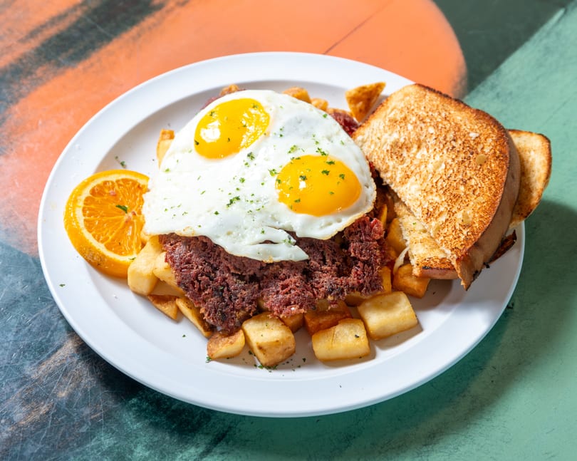 Corned beef hash with two fried eggs, home fries, toast, and an orange slice on a white plate.