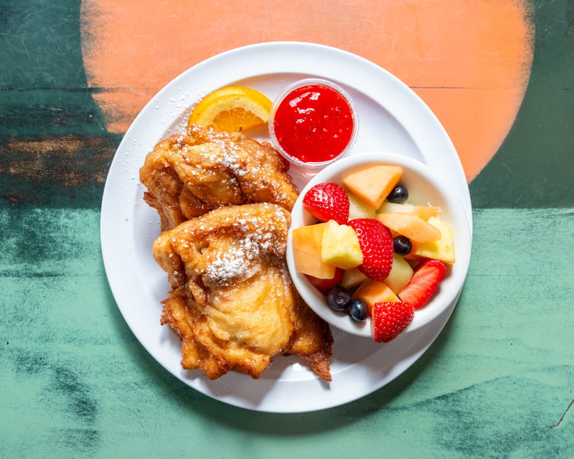 Golden-brown funnel cakes dusted with powdered sugar, served with fresh fruit and strawberry sauce.