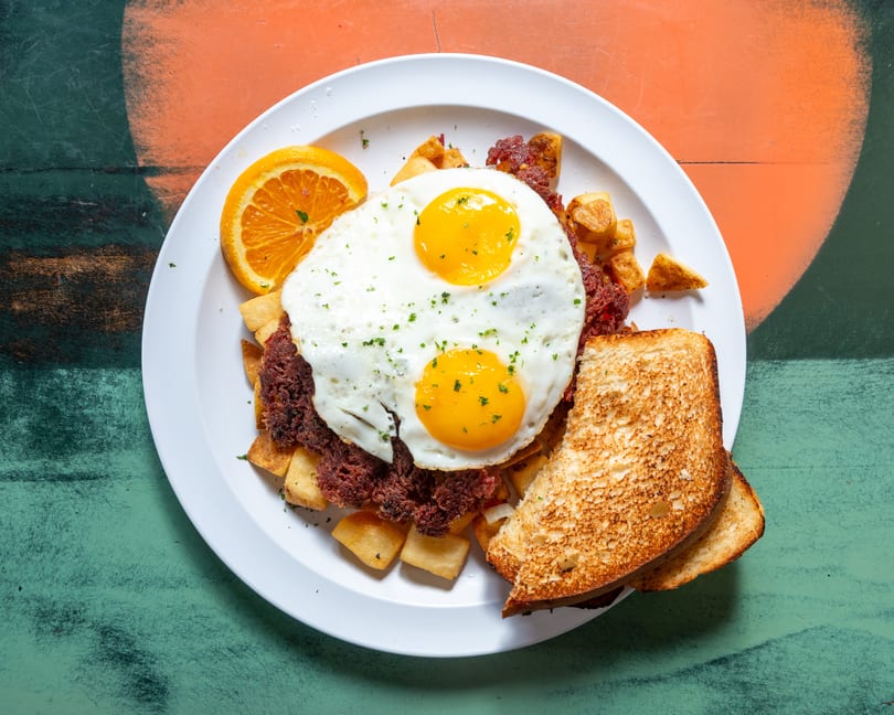 Corned beef hash with two fried eggs, home fries, toast, and an orange slice on a white plate.