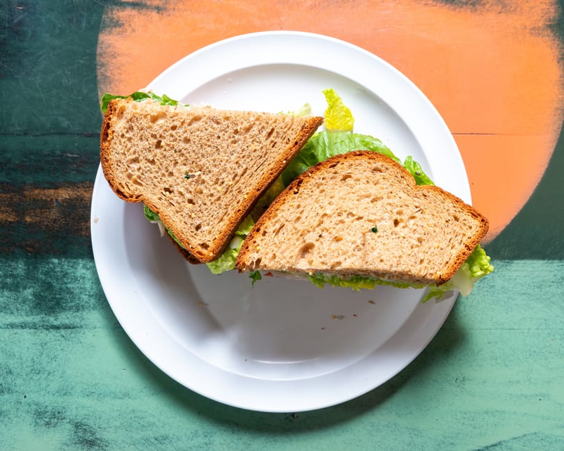 A whole wheat sandwich with lettuce and filling, cut in half, on a white plate.