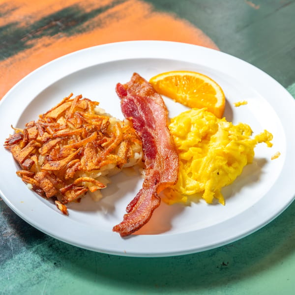 A breakfast plate with crispy hash browns, scrambled eggs, bacon, and an orange slice.