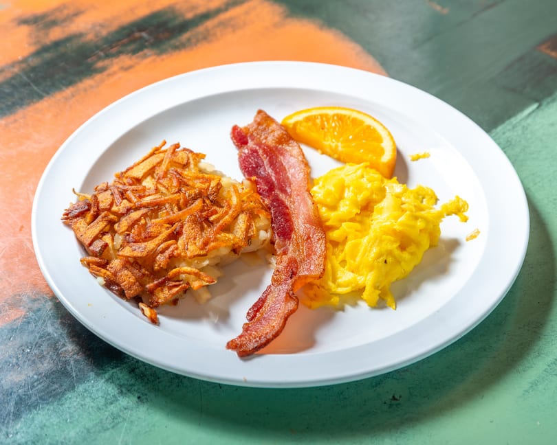 A breakfast plate with crispy hash browns, scrambled eggs, bacon, and an orange slice.