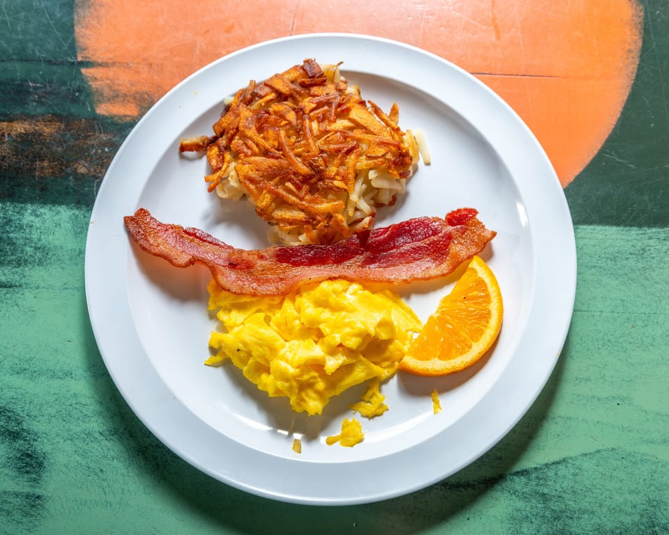 A breakfast plate with crispy hash browns, scrambled eggs, bacon, and an orange slice.
