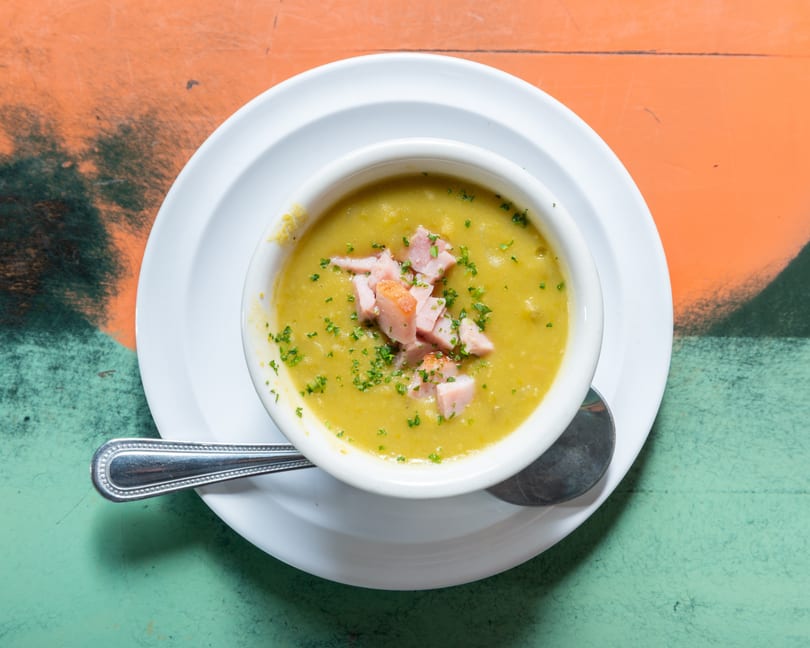 Overhead view of a white bowl of split pea soup with ham and parsley on a colorful table.
