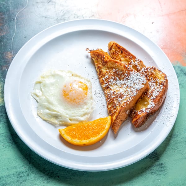 French toast with powdered sugar, a fried egg, and an orange slice on a white plate.