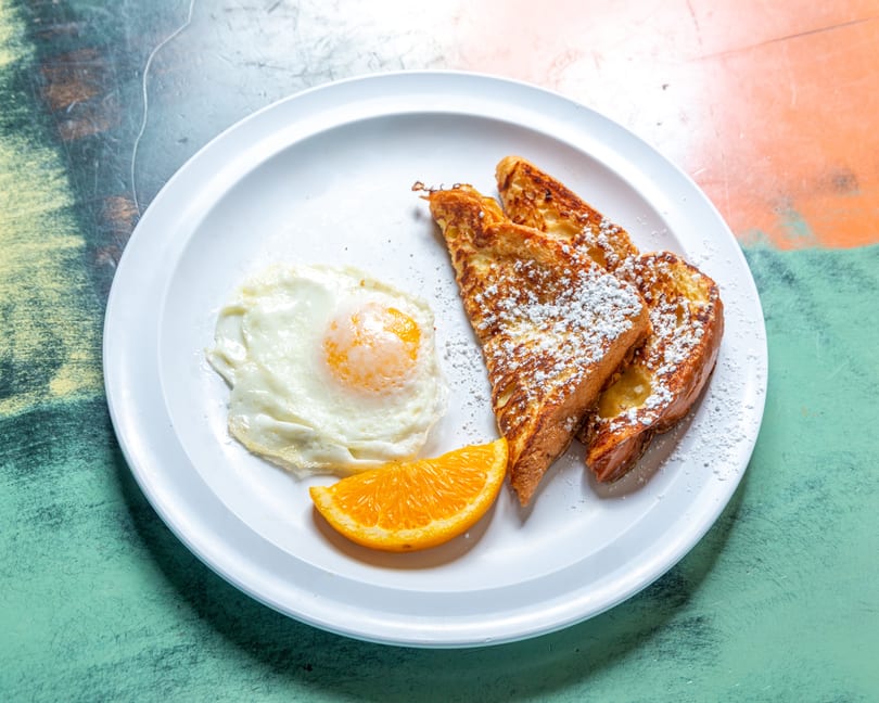 French toast with powdered sugar, a fried egg, and an orange slice on a white plate.