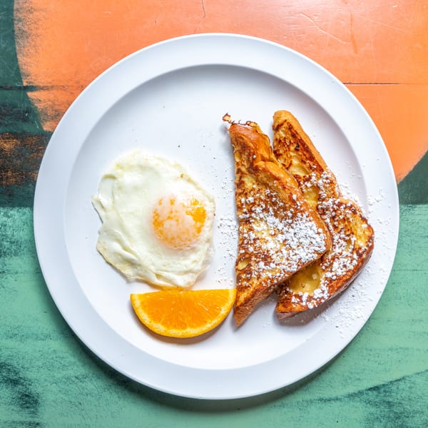 French toast with powdered sugar, a fried egg, and an orange slice on a white plate.