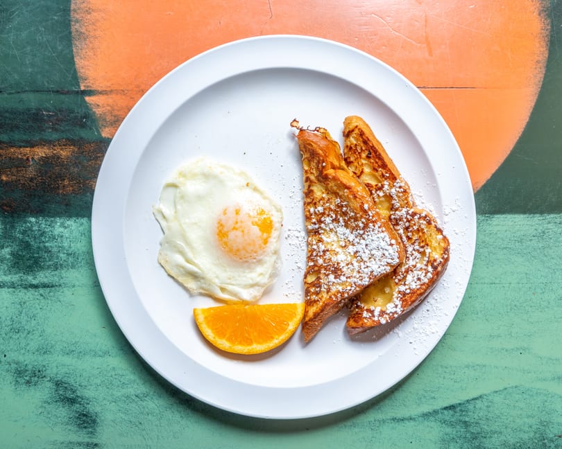 French toast with powdered sugar, a fried egg, and an orange slice on a white plate.