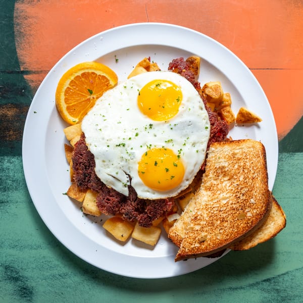 A breakfast plate with two fried eggs, corned beef hash, home fries, toast, and an orange slice.
