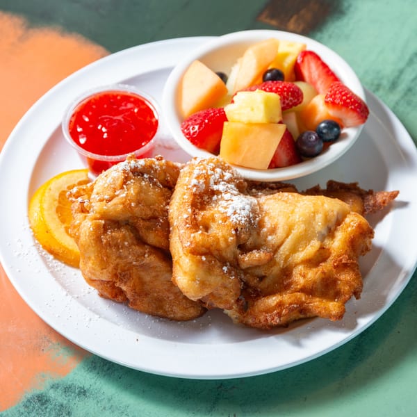 Golden fried fritters dusted with powdered sugar, a side of fruit salad, and strawberry sauce.