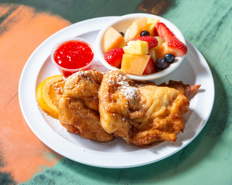 Golden fried fritters dusted with powdered sugar, a side of fruit salad, and strawberry sauce.