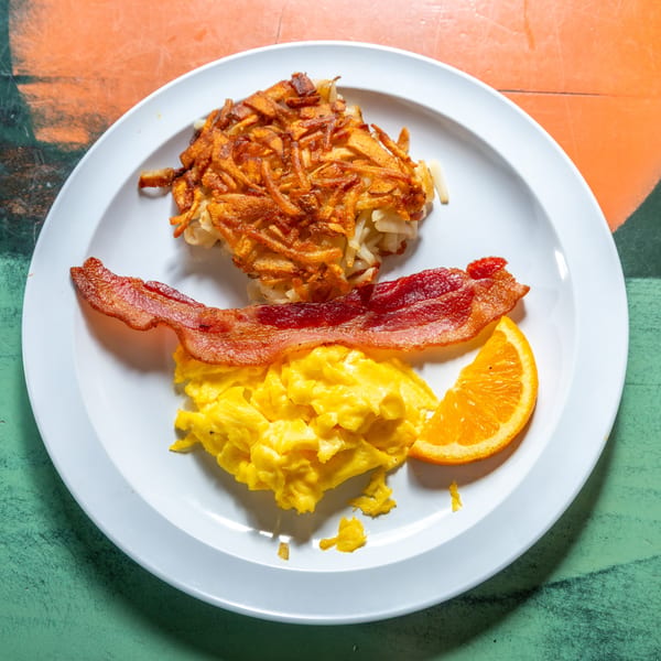 A breakfast plate with crispy hash browns, scrambled eggs, bacon, and an orange slice.