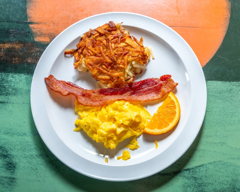 A breakfast plate with crispy hash browns, scrambled eggs, bacon, and an orange slice.