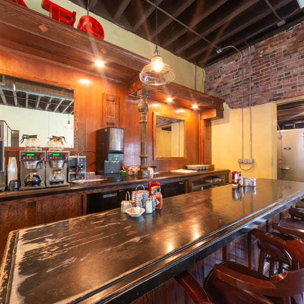 A long wooden bar with stools, coffee machines, and condiments, against a brick and yellow wall.