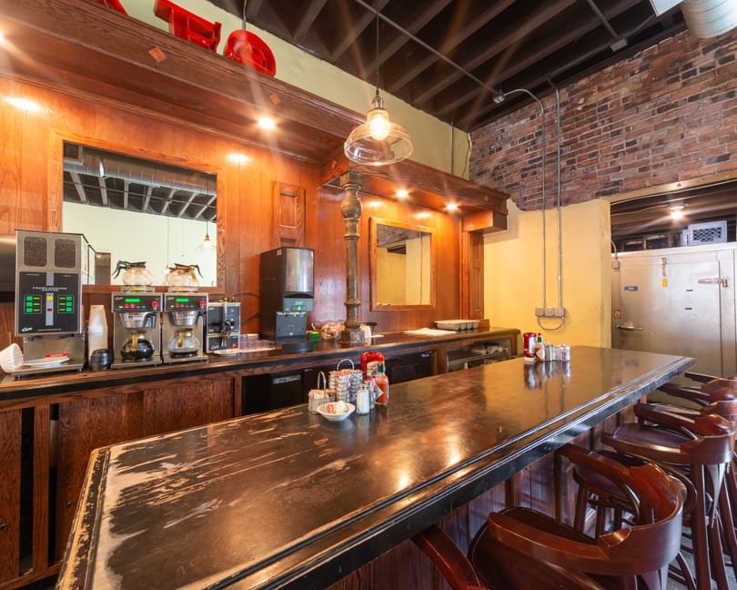 A long wooden bar with stools, coffee machines, and condiments, against a brick and yellow wall.